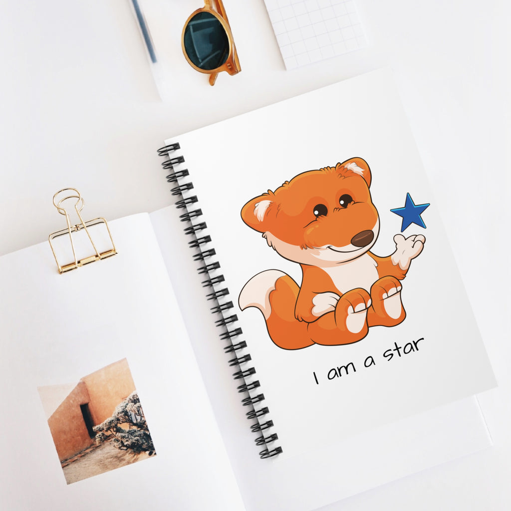 White spiral notebook with a picture of a fox that says I am a star. The notebook is laying closed on a desk.