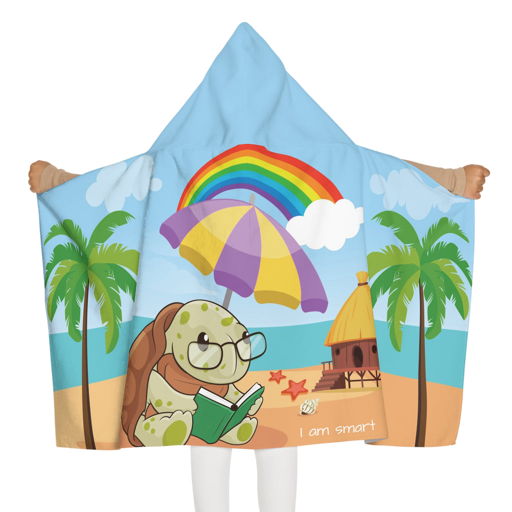 Back-view of a girl wearing a hooded towel and holding it open. The towel has a scene of a turtle reading a book under an umbrella on the beach, a rainbow in the background, and the phrase "I am smart" along the bottom.