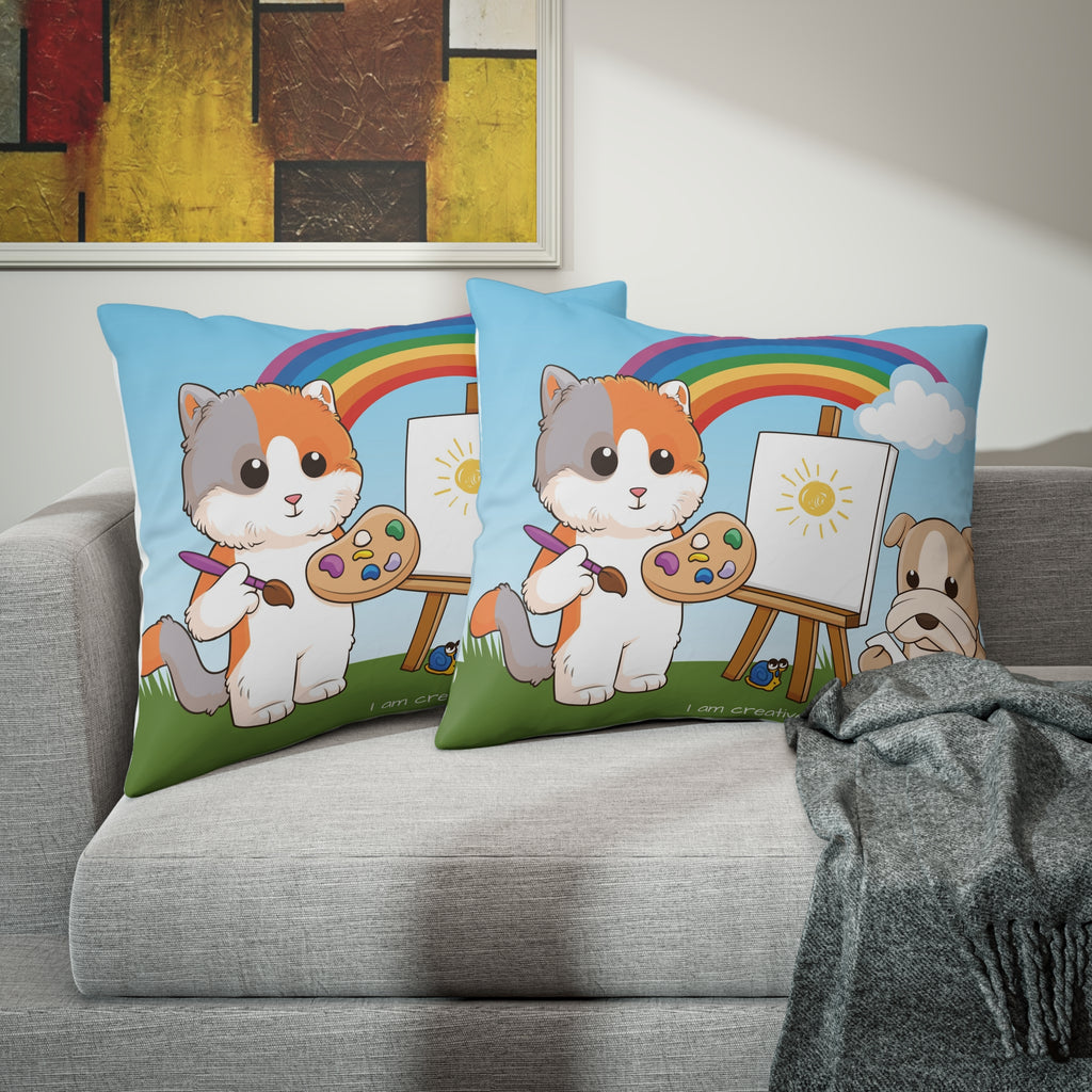Two pillows sitting on a grey couch. The pillows have on pillowcases with a scene of a cat painting on a canvas next to a dog, a rainbow in the background, and the phrase "I am creative" along the bottom.
