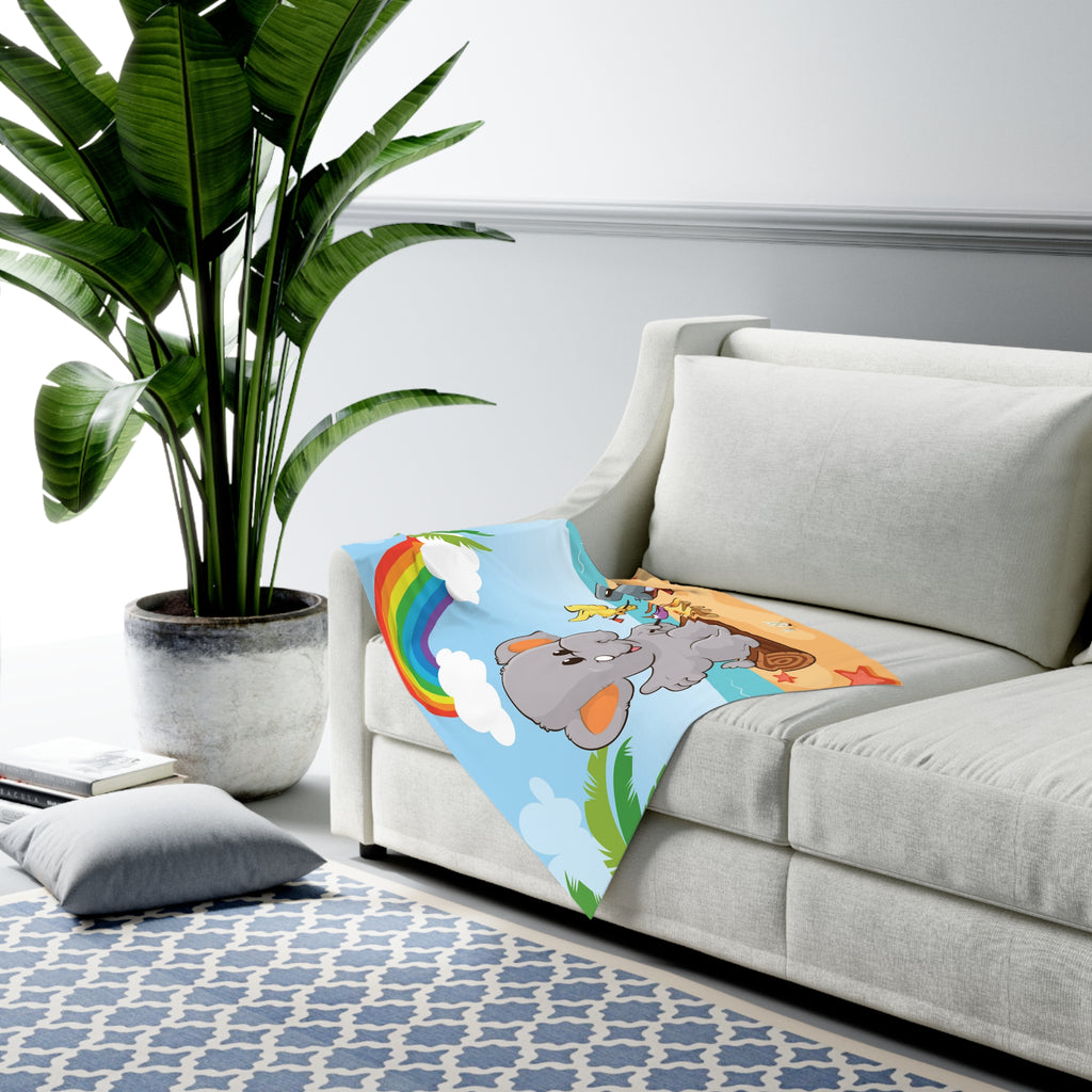 Full-color swaddle blanket with a scene of an elephant having a bonfire with a bird and fish on the beach, a rainbow in the background, and the phrase "I am calm" along the bottom. The blanket is draped over the armrest of a couch.