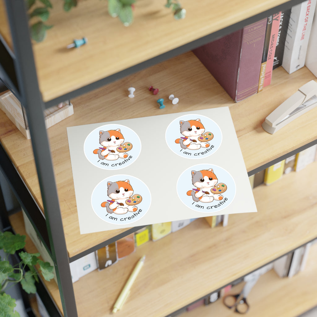 A sheet of 4 round stickers with a picture of a cat that says I am creative. The sticker sheet sits on a shelf of a bookcase.