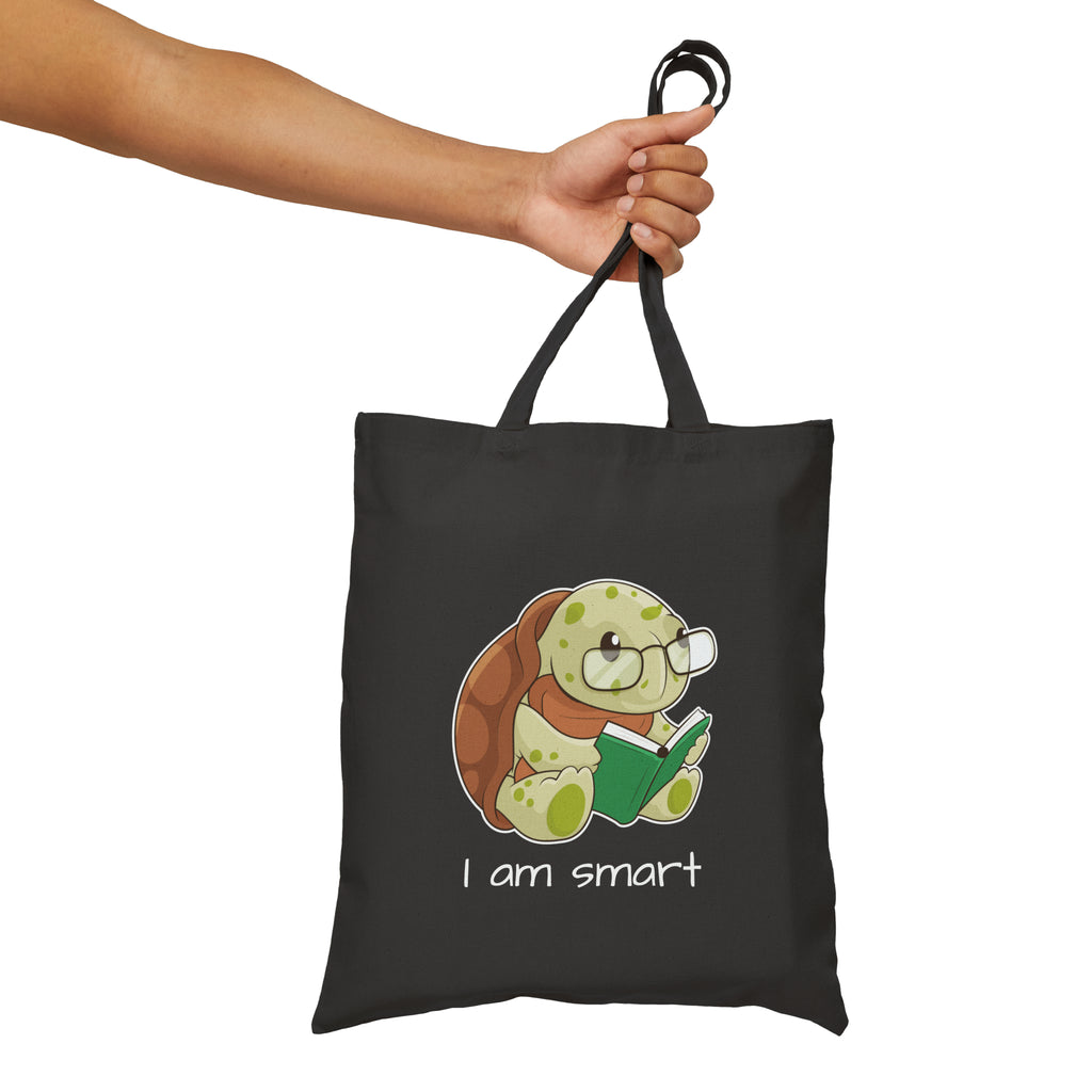 A hand holding a black tote bag with a picture of a turtle that says I am smart.