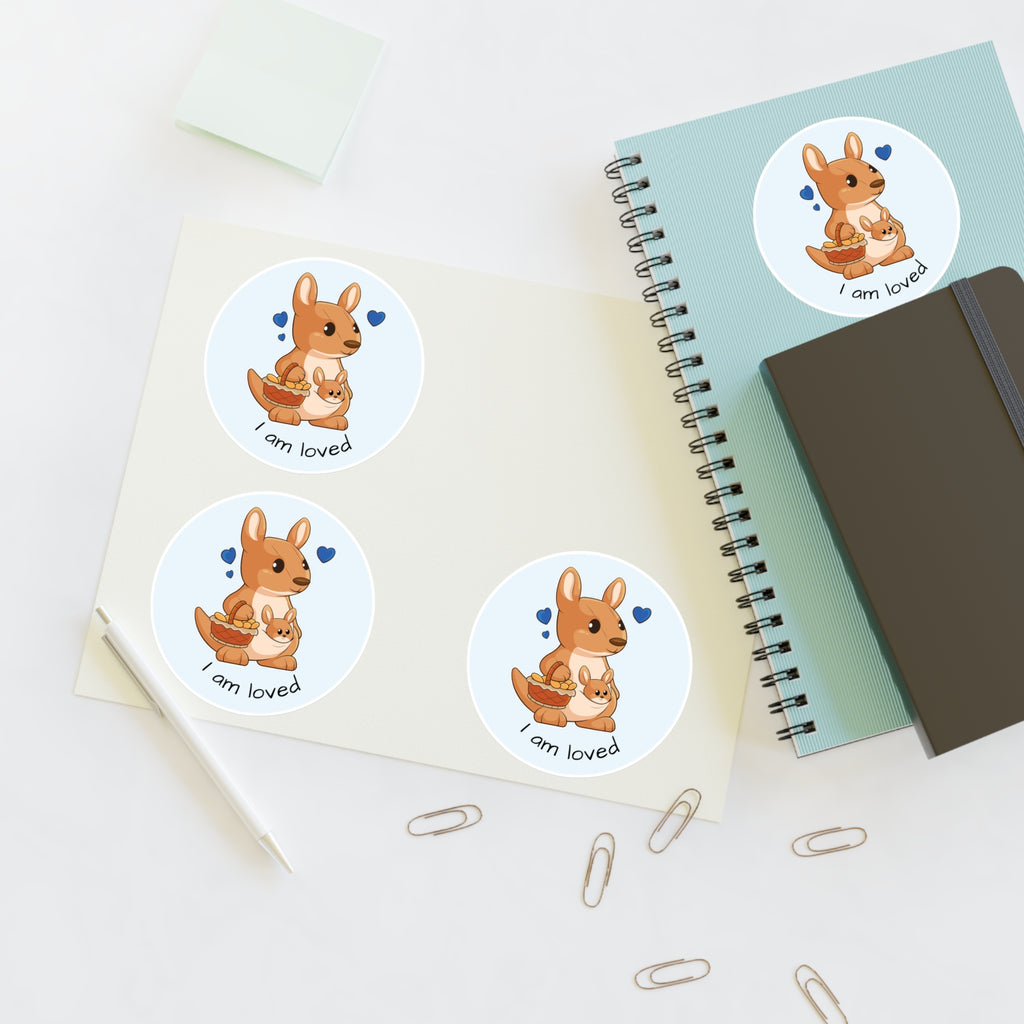 A sheet of 3 round stickers with a picture of a kangaroo that says I am loved. The sticker sheet sits on a table next to a notebook with the fourth sticker on it.
