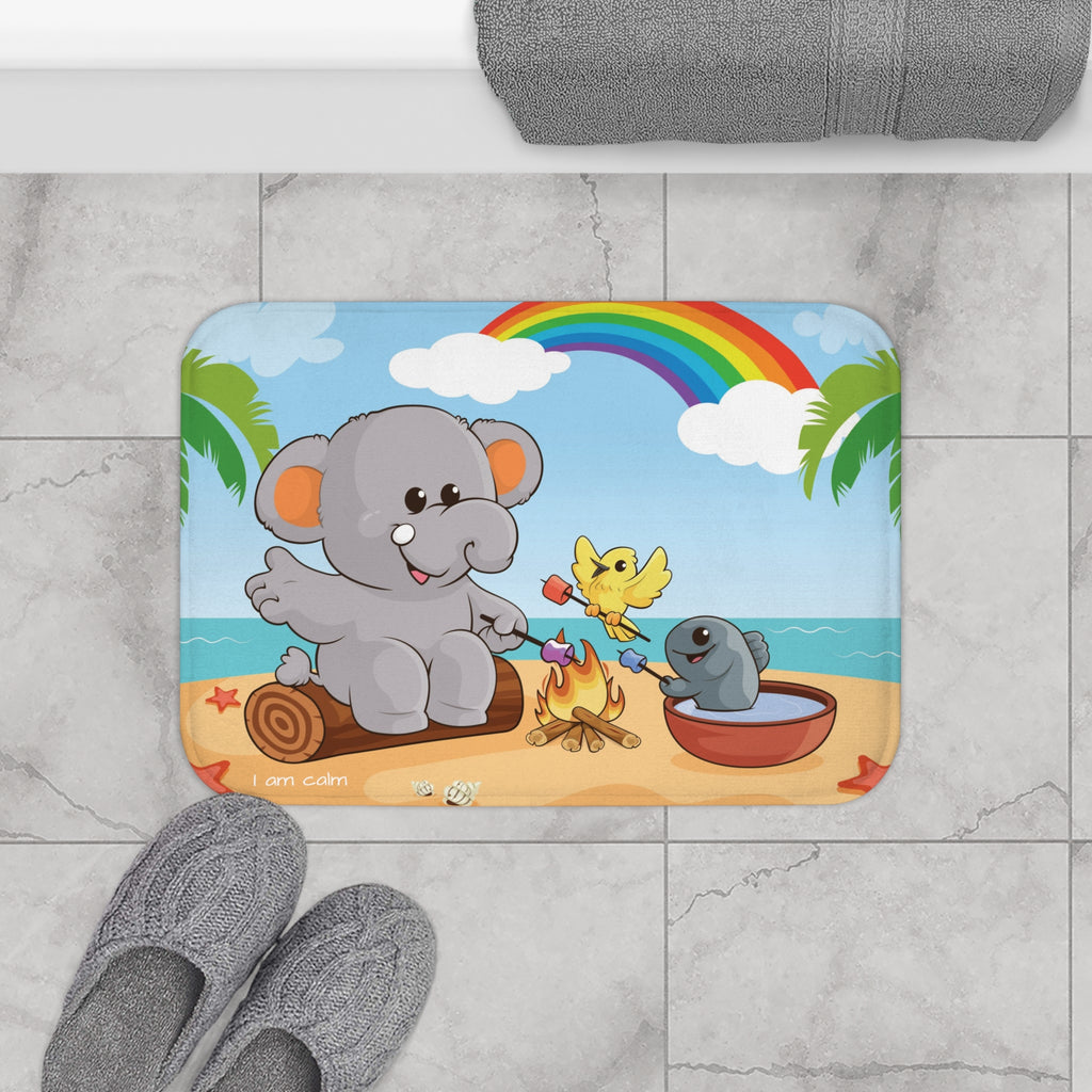 A 24 by 17 inch bath mat on the tiled floor of a bathroom. The bath mat has a scene of an elephant having a bonfire with a bird and fish on the beach, a rainbow in the background, and the phrase "I am calm" along the bottom.