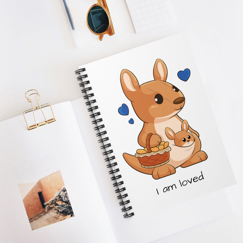 White spiral notebook with a picture of a kangaroo that says I am loved. The notebook is laying closed on a desk.