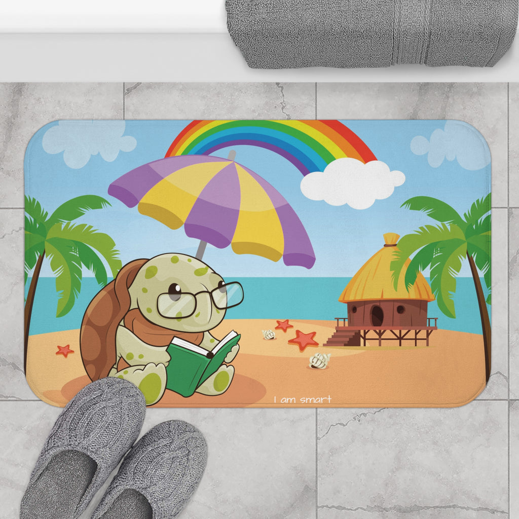 A 34 by 21 inch bath mat on the tiled floor of a bathroom. The bath mat has a scene of a turtle reading under an umbrella on a beach with a rainbow in the background and the phrase "I am smart" along the bottom.