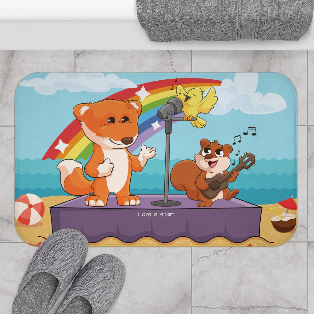 A 34 by 21 inch bath mat on the tiled floor of a bathroom. The bath mat has a scene of a fox singing with a bird and squirrel on a stage on the beach with a rainbow in the background and the phrase "I am a star" along the bottom.
