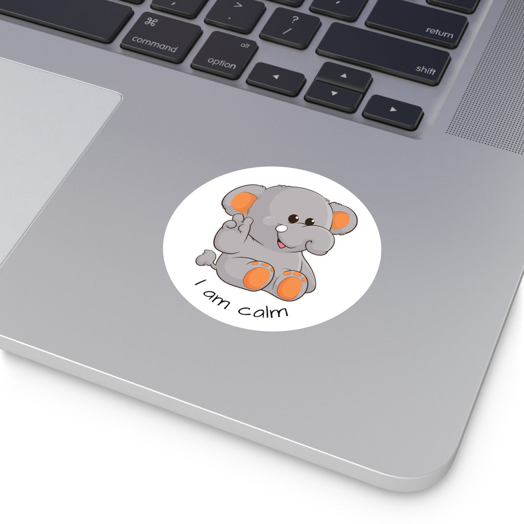 A 3 by 3 inch round white vinyl sticker with a picture of an elephant that says I am calm. The sticker is on a grey laptop next to the mousepad.
