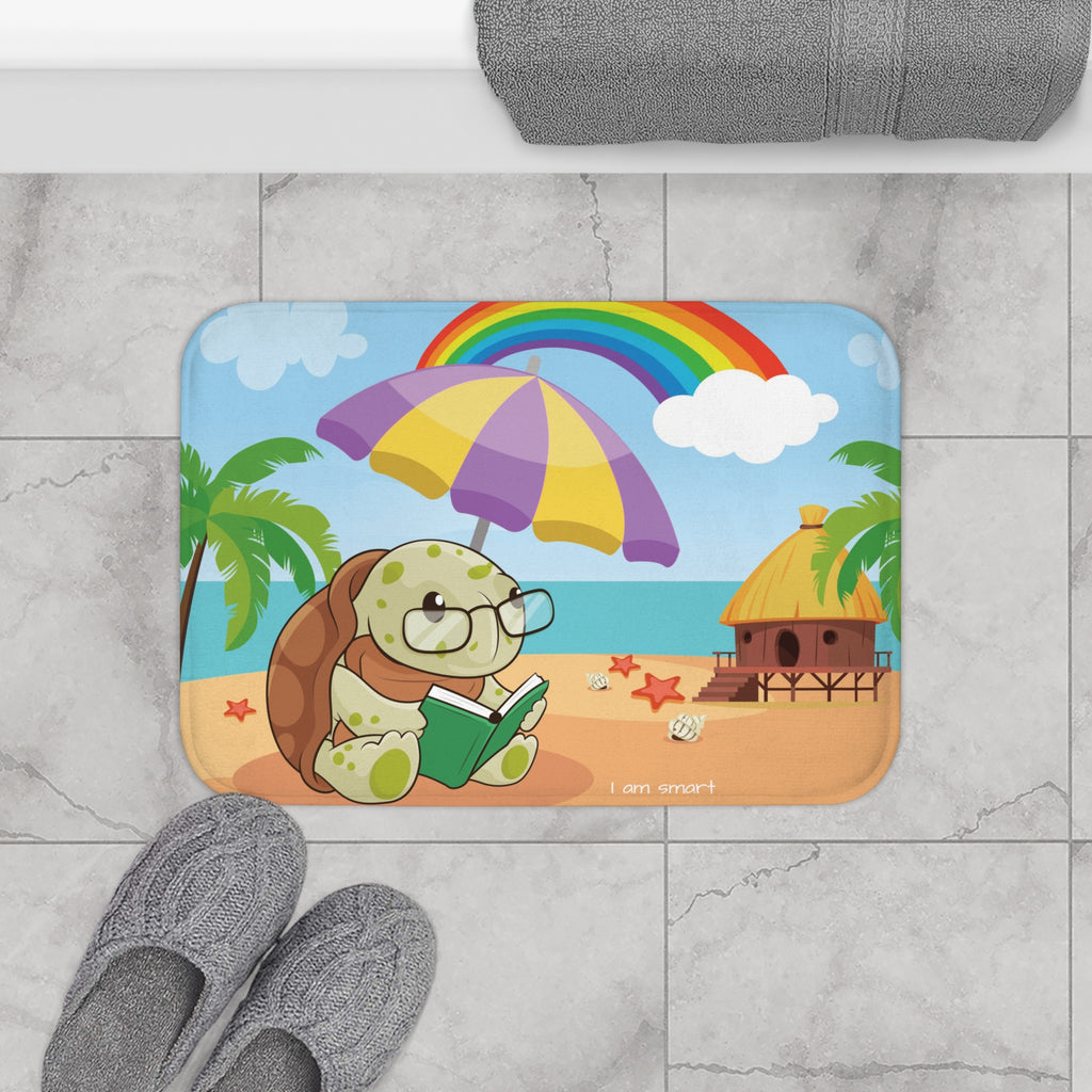 A 24 by 17 inch bath mat on the tiled floor of a bathroom. The bath mat has a scene of a turtle reading under an umbrella on a beach with a rainbow in the background and the phrase "I am smart" along the bottom.