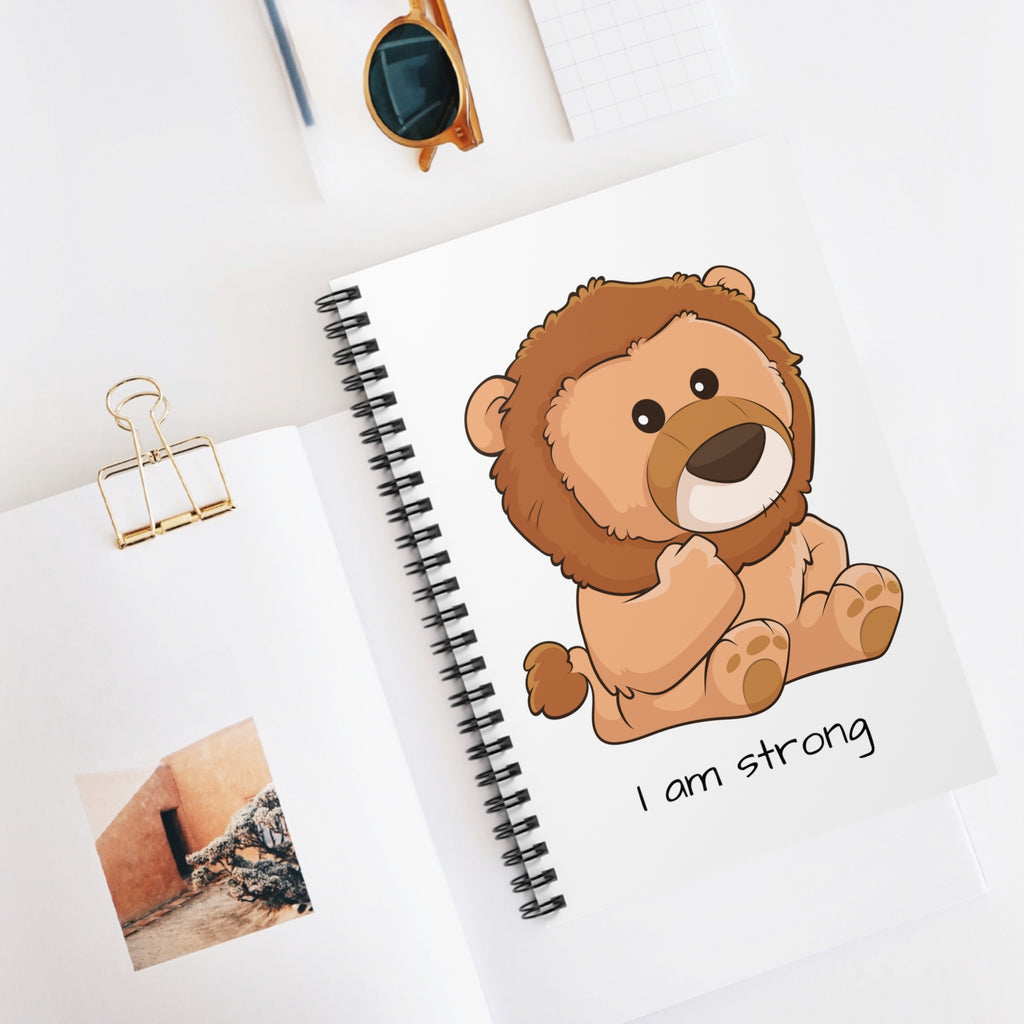 White spiral notebook with a picture of a lion that says I am strong. The notebook is laying closed on a desk.