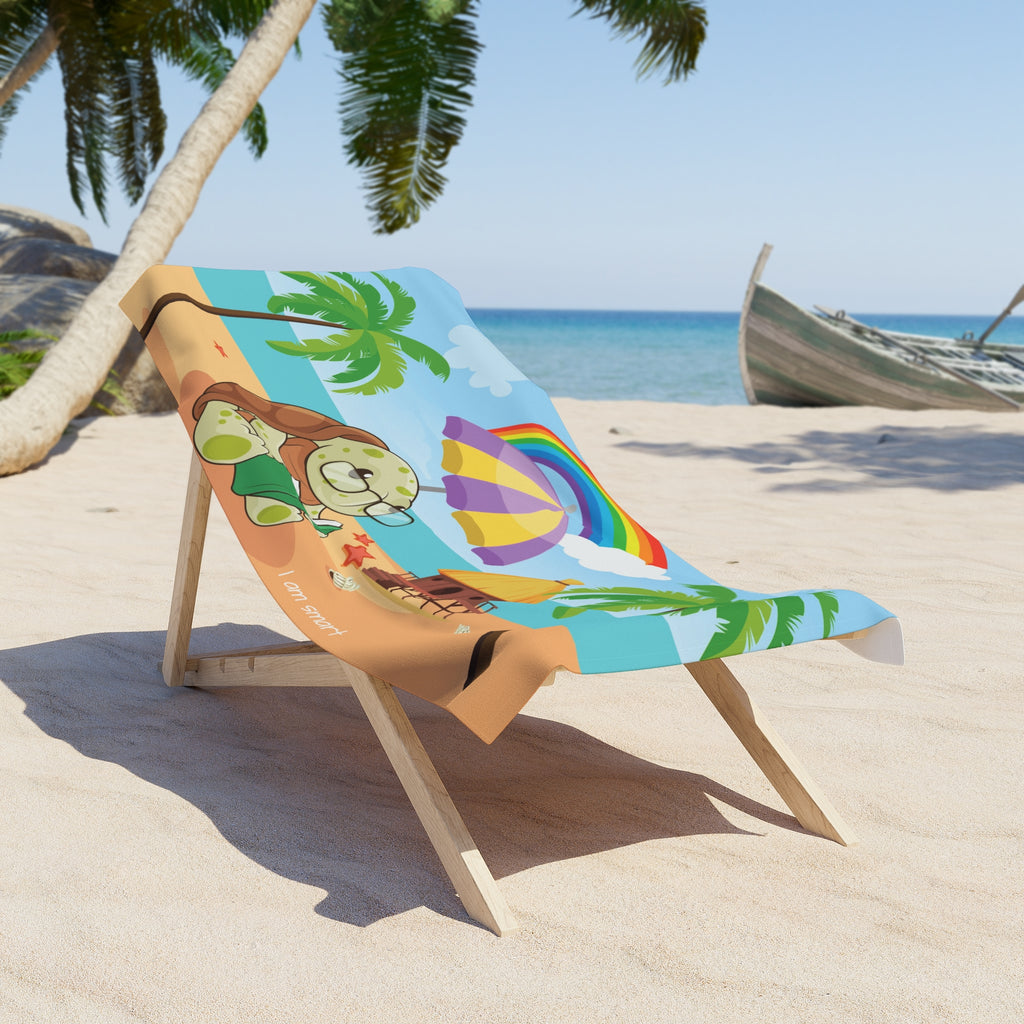 A 30 by 60 inch beach towel draped over a chair on a beach. The towel has a scene of a turtle reading a book under an umbrella on the beach, a rainbow in the background, and the phrase "I am smart" along the bottom.