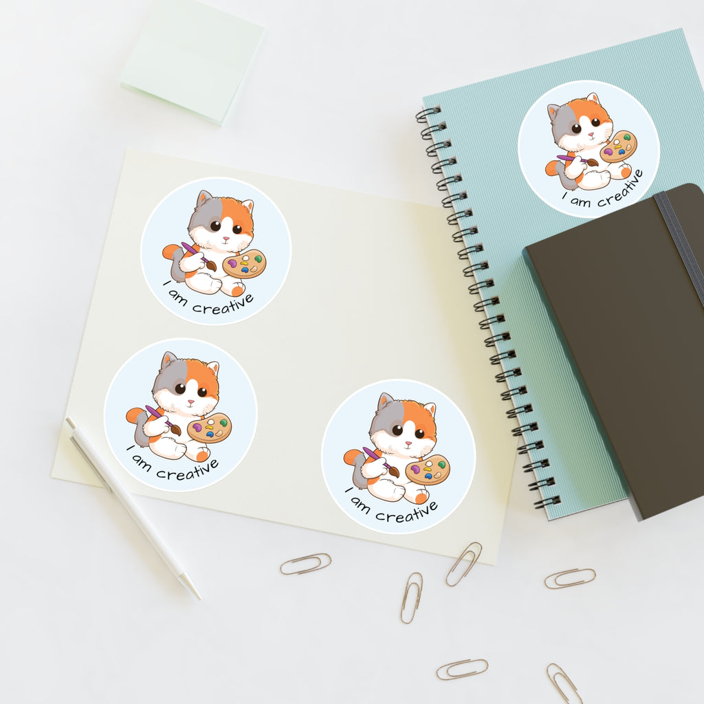 A sheet of 3 round stickers with a picture of a cat that says I am creative. The sticker sheet sits on a table next to a notebook with the fourth sticker on it.