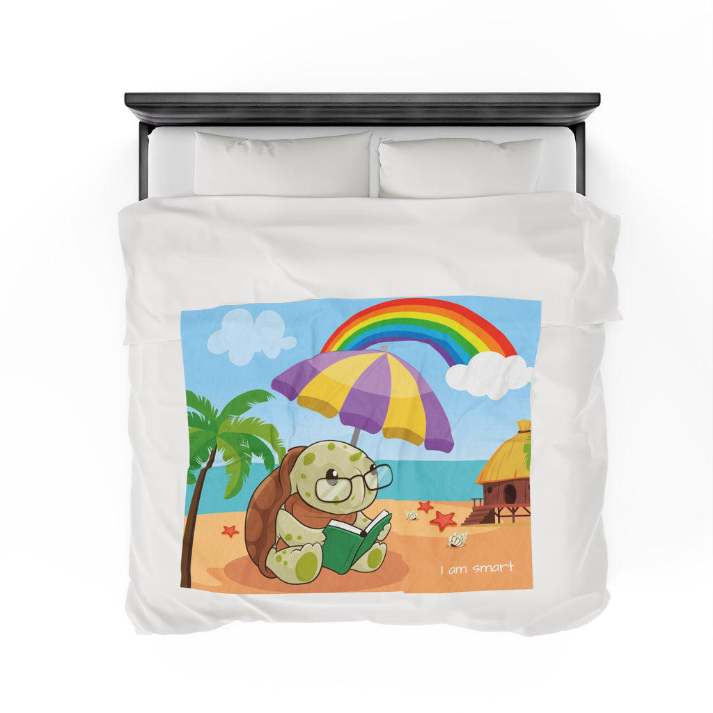 Top-view of a 50 by 60 inch blanket on a queen-sized bed. The blanket has a scene of a turtle reading under an umbrella on the beach, a rainbow in the background, and the phrase "I am smart" along the bottom.