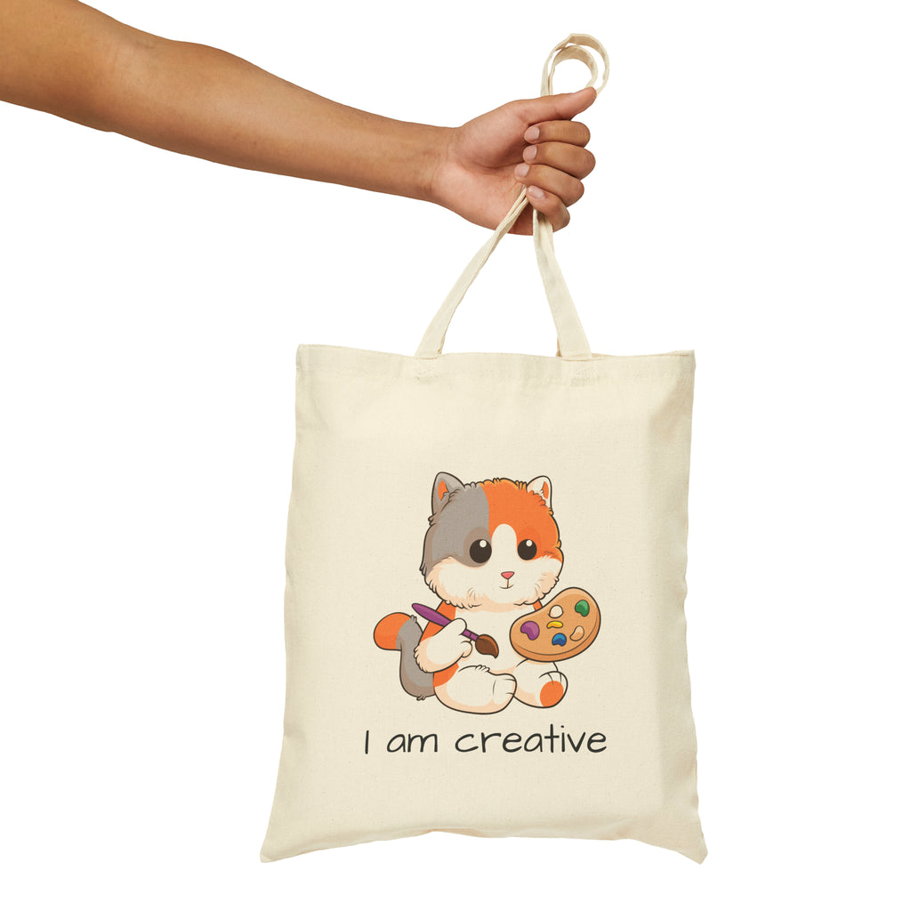 A hand holding a natural tan tote bag with a picture of a cat that says I am creative.