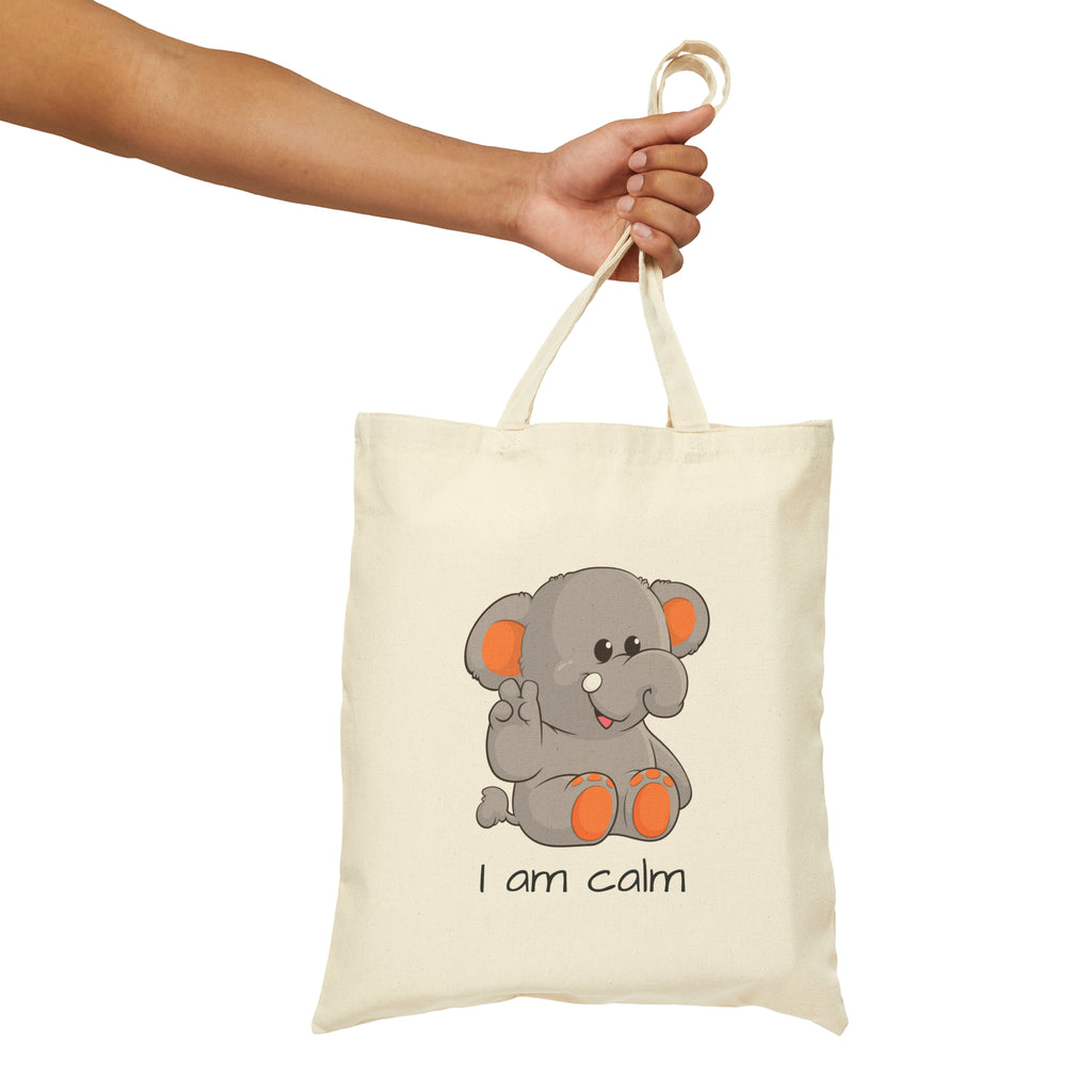 A hand holding a natural tan tote bag with a picture of an elephant that says I am calm.