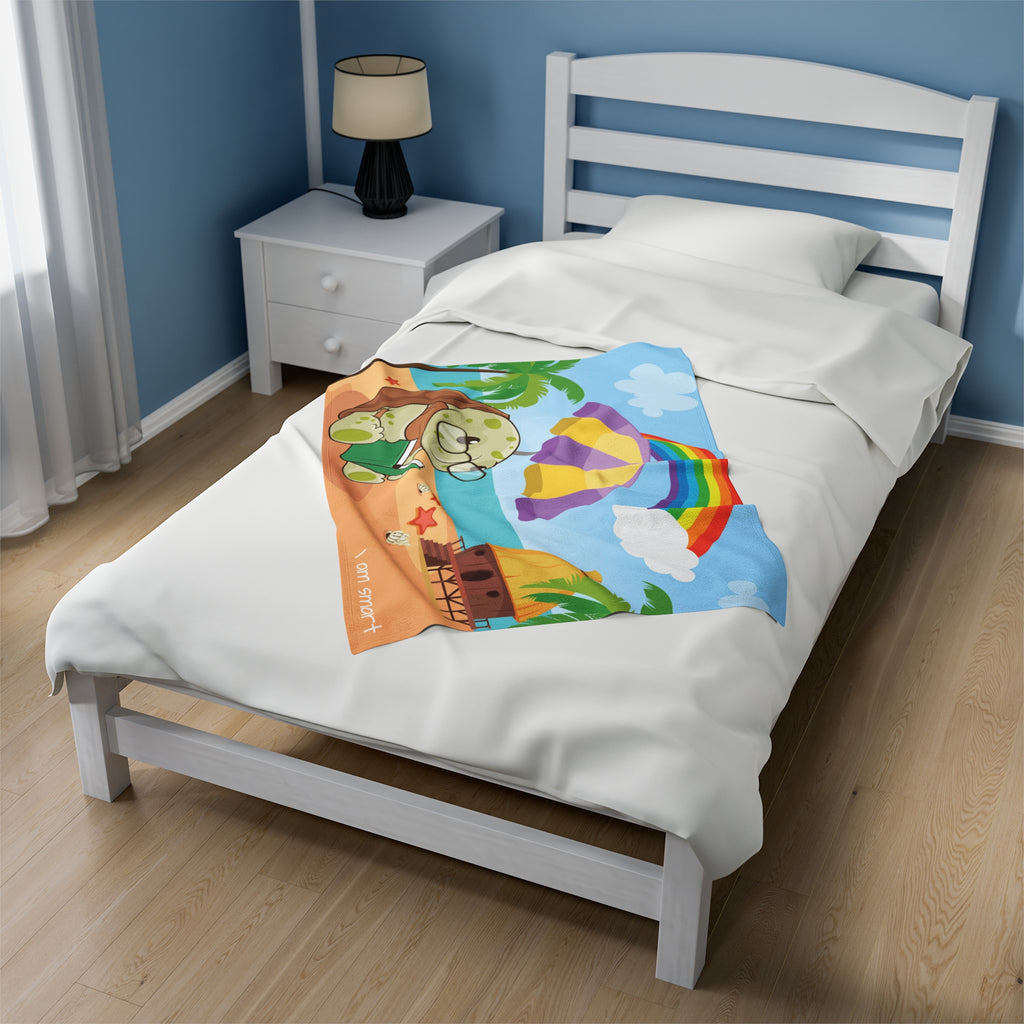 A 30 by 40 inch blanket on a twin-sized bed in a bedroom. The blanket has a scene of a turtle reading under an umbrella on the beach, a rainbow in the background, and the phrase "I am smart" along the bottom.
