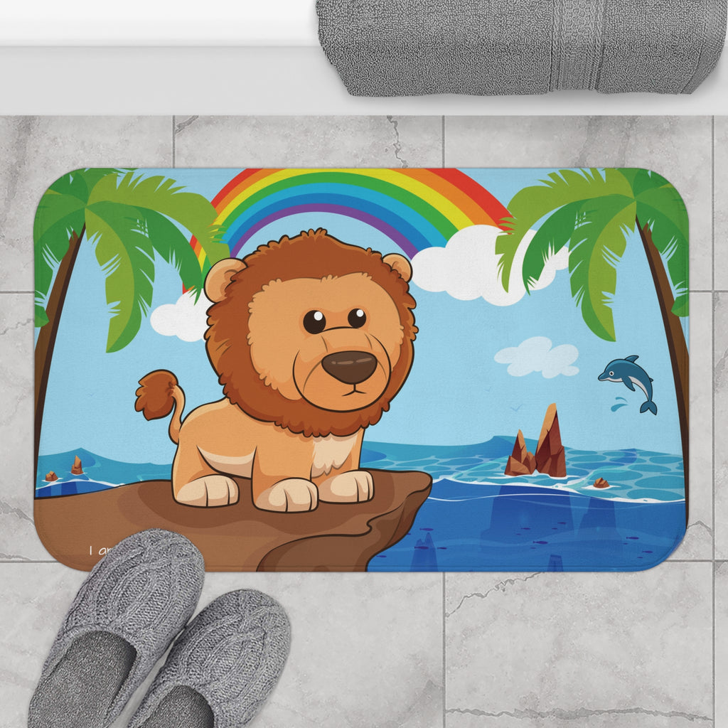A 34 by 21 inch bath mat on the tiled floor of a bathroom. The bath mat has a scene of a lion standing on a cliff over the ocean with a rainbow in the background and the phrase "I am strong" along the bottom.