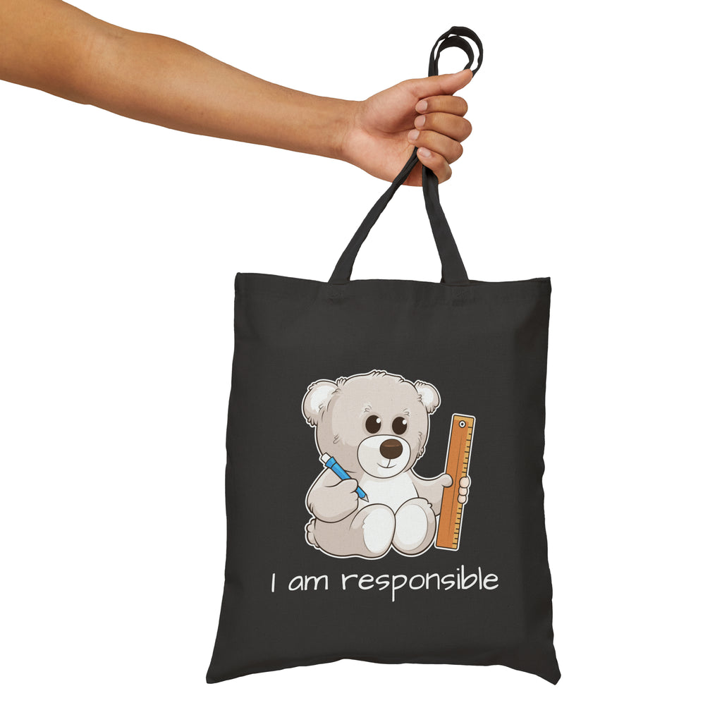 A hand holding a black tote bag with a picture of a bear that says I am responsible.
