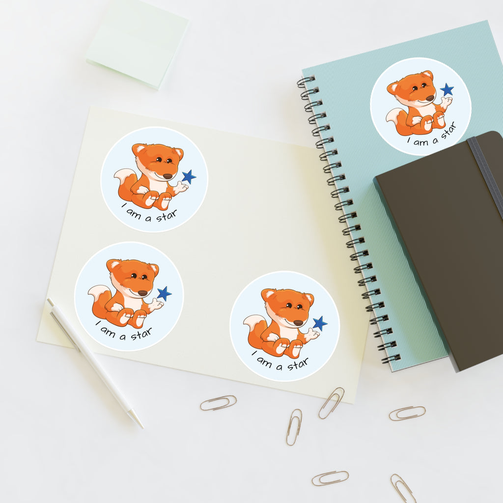 A sheet of 3 round stickers with a picture of a fox that says I am a star. The sticker sheet sits on a table next to a notebook with the fourth sticker on it.