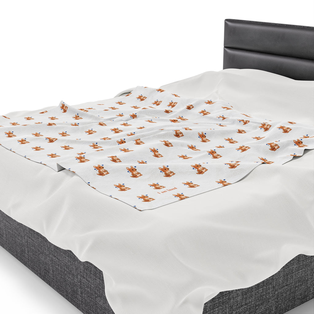 Side-view of a 50 by 60 inch blanket on a queen-sized bed. The blanket has a repeating pattern of a kangaroo and the phrase “I am loved” in the bottom left corner.