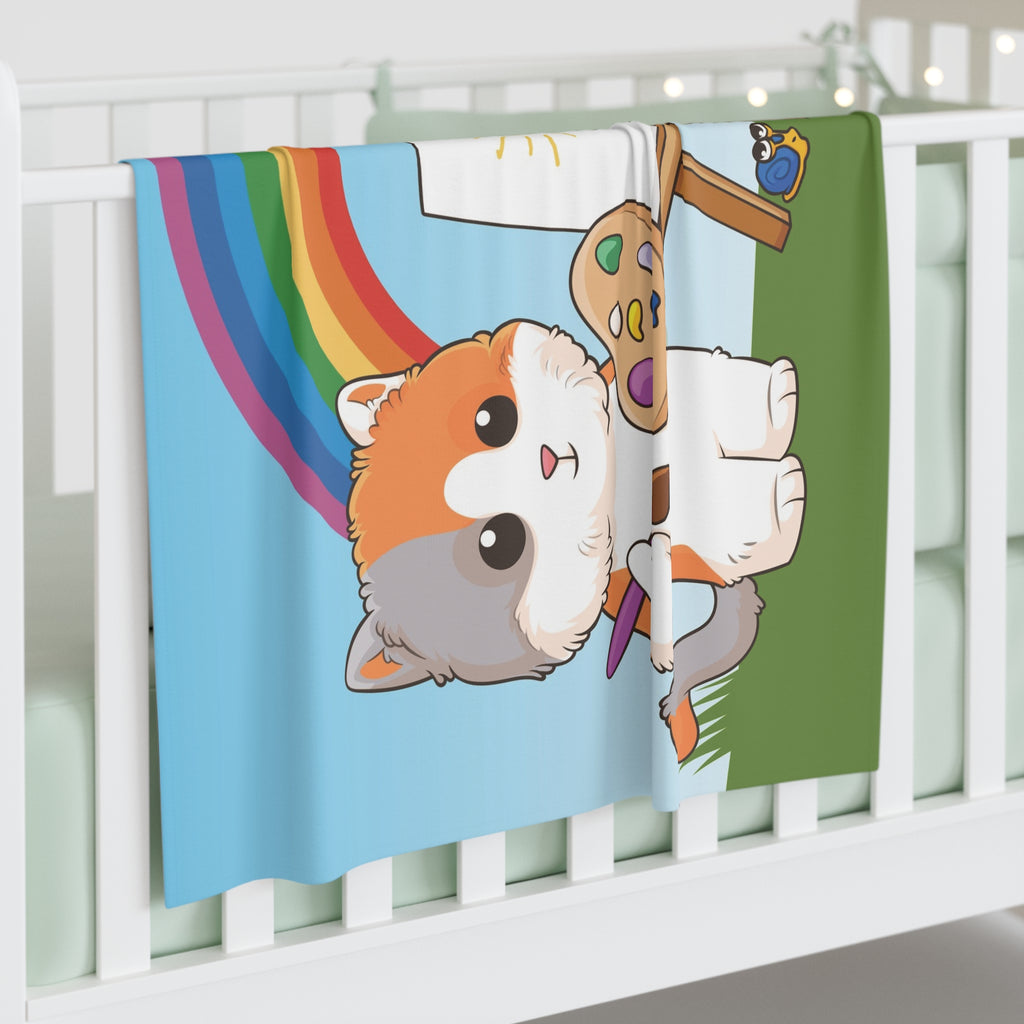 Full-color swaddle blanket with a cat painting on a canvas next to a dog and a rainbow in the background. The blanket is draped over the side of a baby crib.