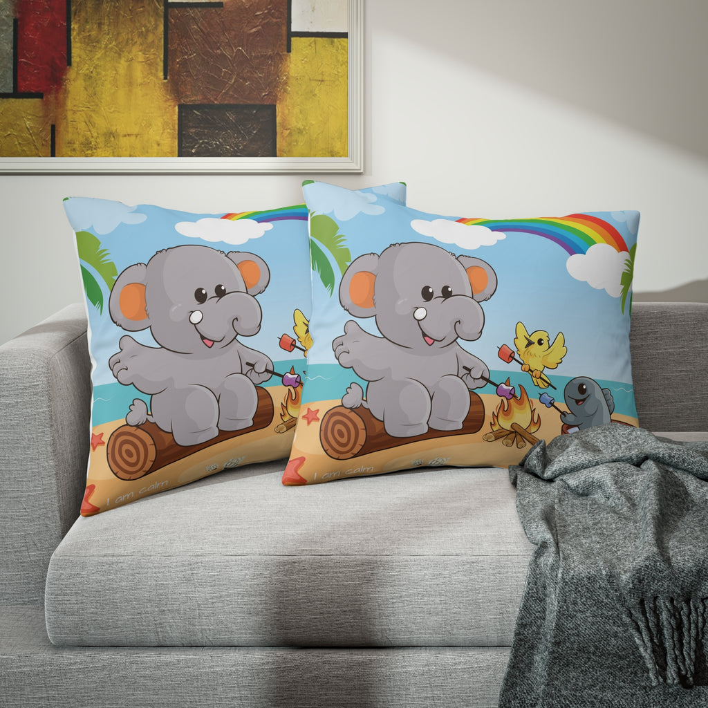 Two pillows sitting on a grey couch. The pillows have on pillowcases with a scene of an elephant having a bonfire with a bird and fish on the beach, a rainbow in the background, and the phrase "I am calm" along the bottom.