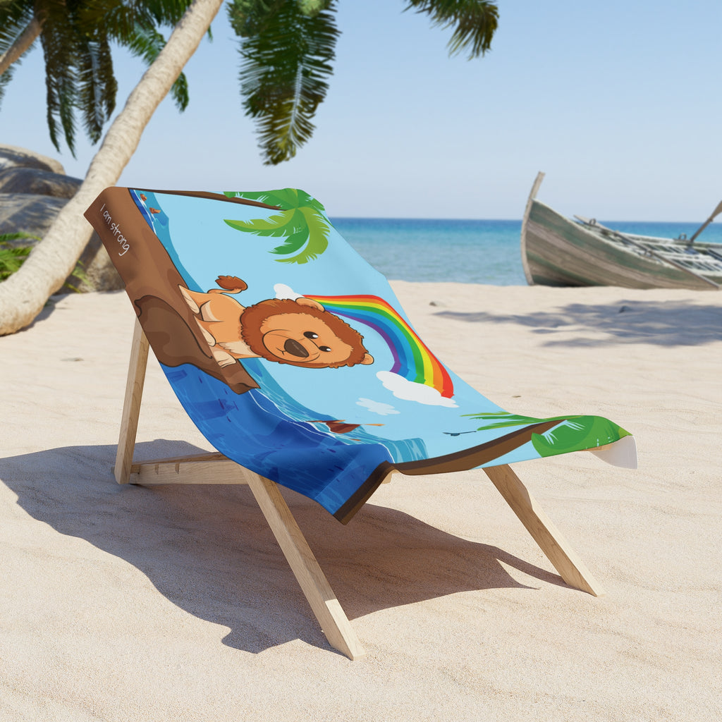 A 30 by 60 inch beach towel draped over a chair on a beach. The towel has a scene of a lion standing on a cliff over the ocean, a rainbow in the background, and the phrase "I am strong" along the bottom.