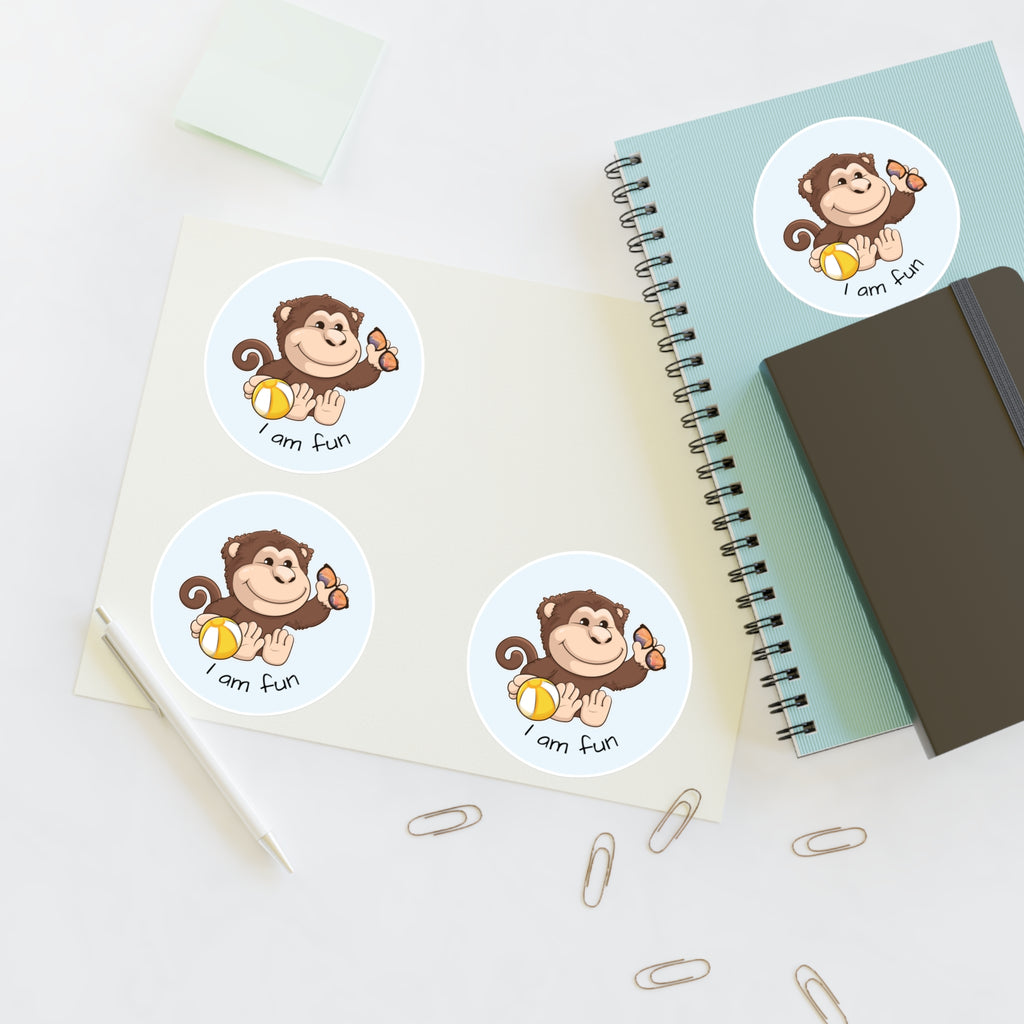 A sheet of 3 round stickers with a picture of a monkey that says I am fun. The sticker sheet sits on a table next to a notebook with the fourth sticker on it.