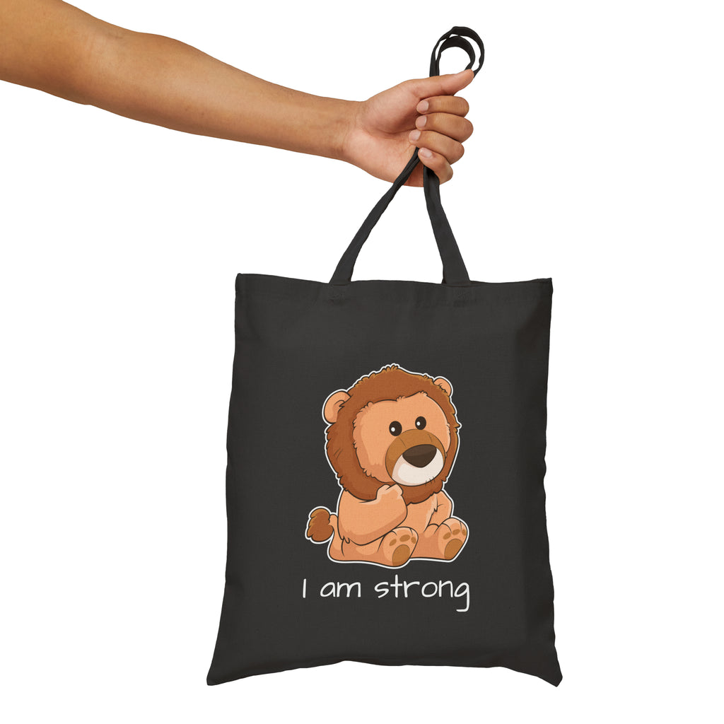 A hand holding a black tote bag with a picture of a lion that says I am strong.