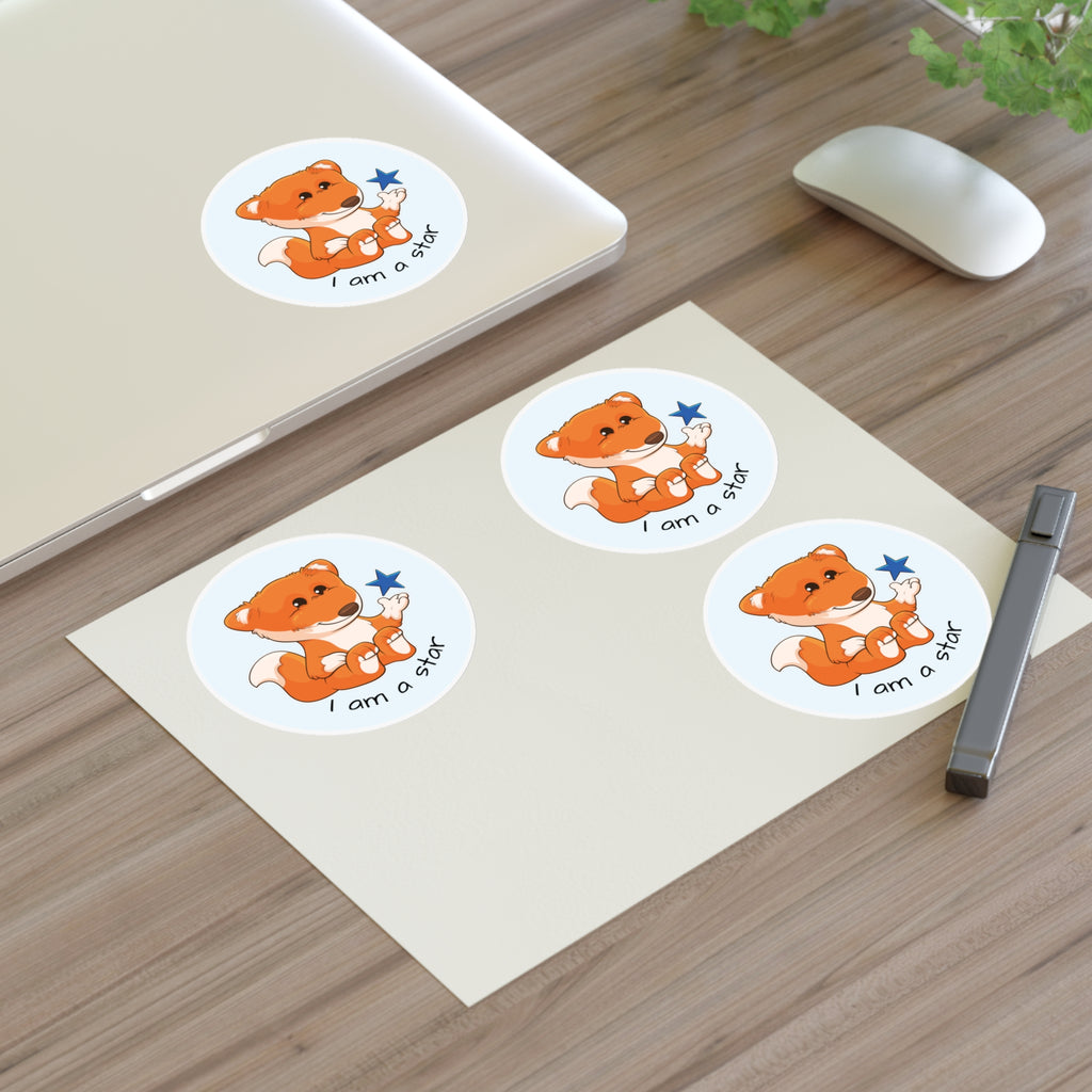 A sheet of 3 round stickers with a picture of a fox that says I am a star. The sticker sheet sits on a table next to a laptop with the fourth sticker on it.