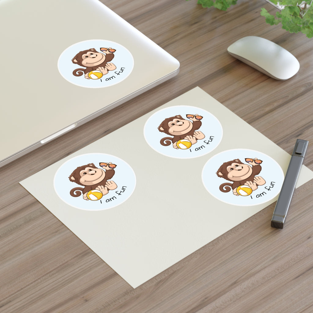 A sheet of 3 round stickers with a picture of a monkey that says I am fun. The sticker sheet sits on a table next to a laptop with the fourth sticker on it.