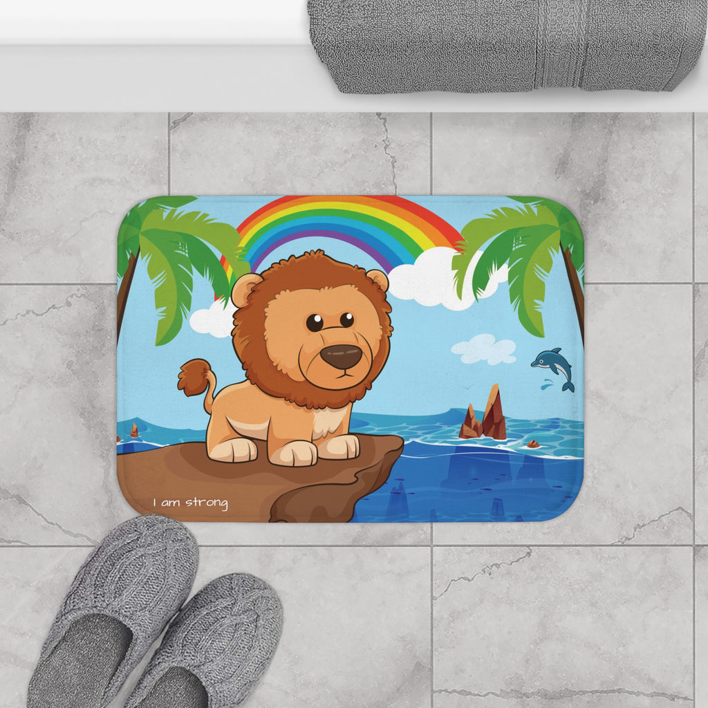 A 24 by 17 inch bath mat on the tiled floor of a bathroom. The bath mat has a scene of a lion standing on a cliff over the ocean with a rainbow in the background and the phrase "I am strong" along the bottom.