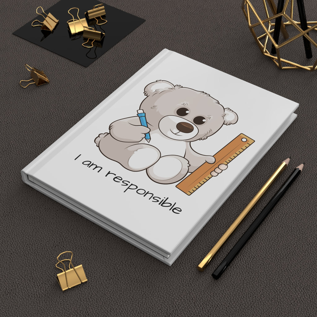 White hardcover journal with a picture of a bear that says I am responsible. The journal is laying closed on a desk.