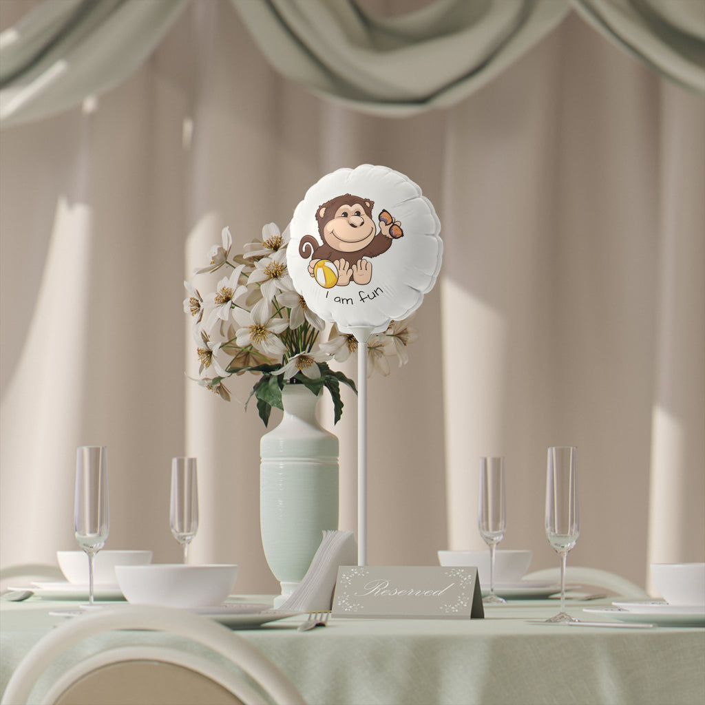 A round white mylar balloon on a stick with a picture of a monkey that says I am fun. The balloon sits on a table decorated for an event.