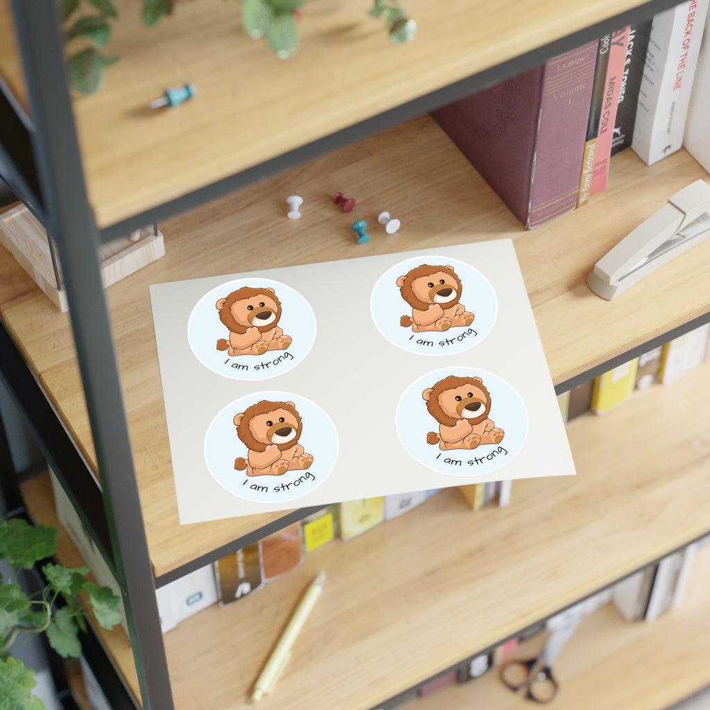 A sheet of 4 round stickers with a picture of a lion that says I am strong. The sticker sheet sits on a shelf of a bookcase.