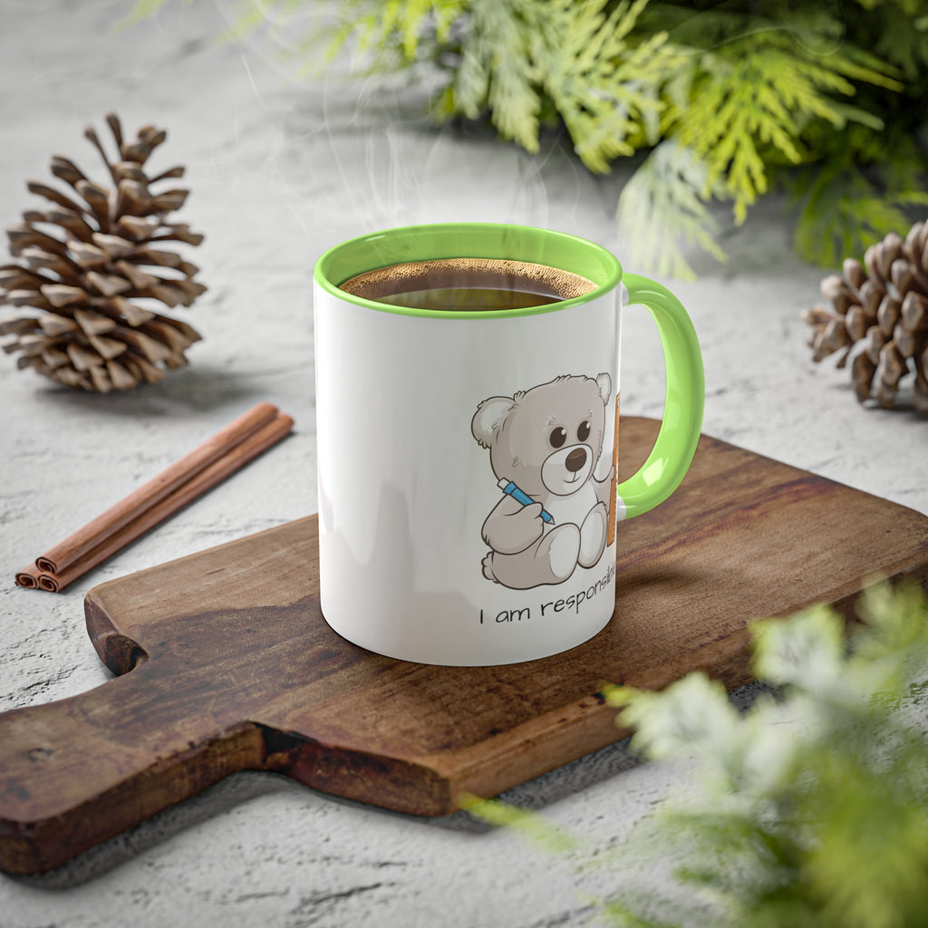 A white mug with a light green handle and interior and a picture of a bear that says I am responsible. The mug has hot coffee in it and sits on a wood cutting board surrounded by pinecones and pine trees.