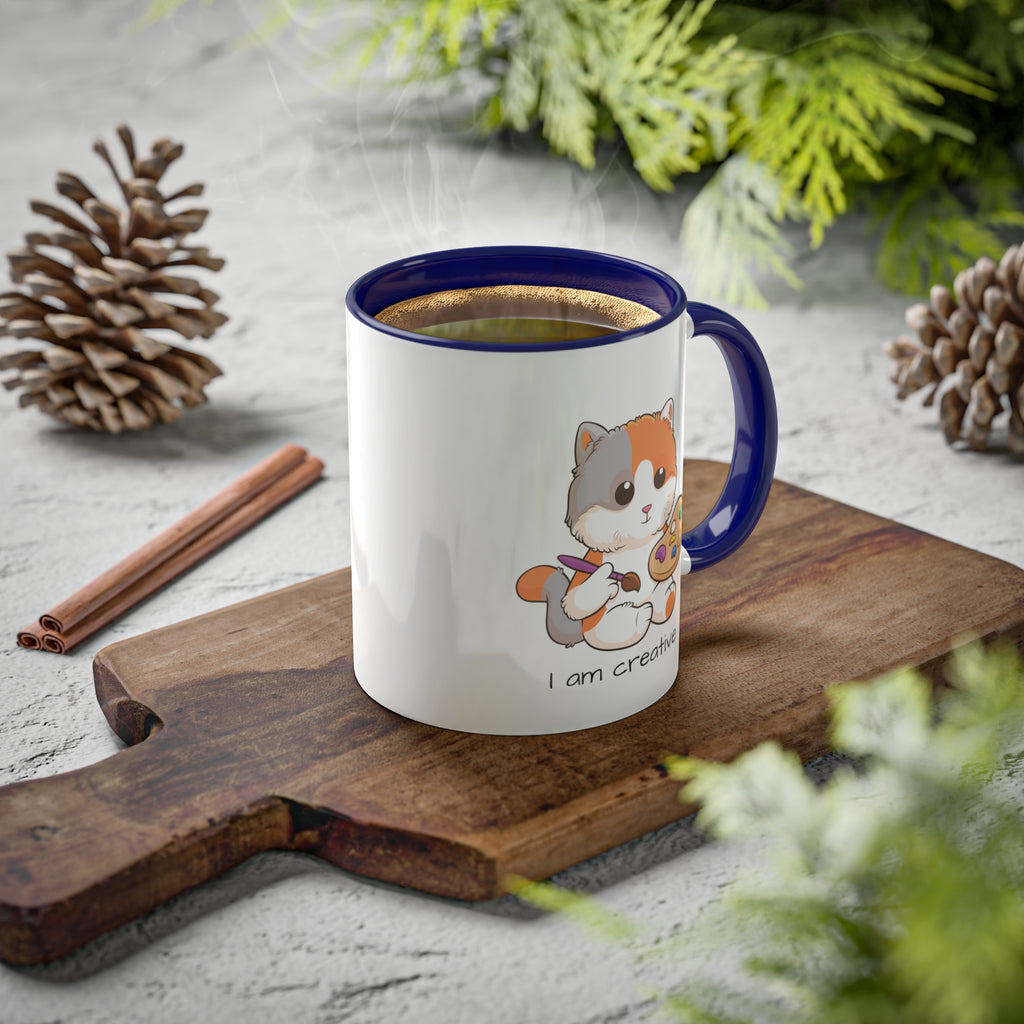 A white mug with a dark blue handle and interior and a picture of a cat that says I am creative. The mug has hot coffee in it and sits on a wood cutting board surrounded by pinecones and pine trees.