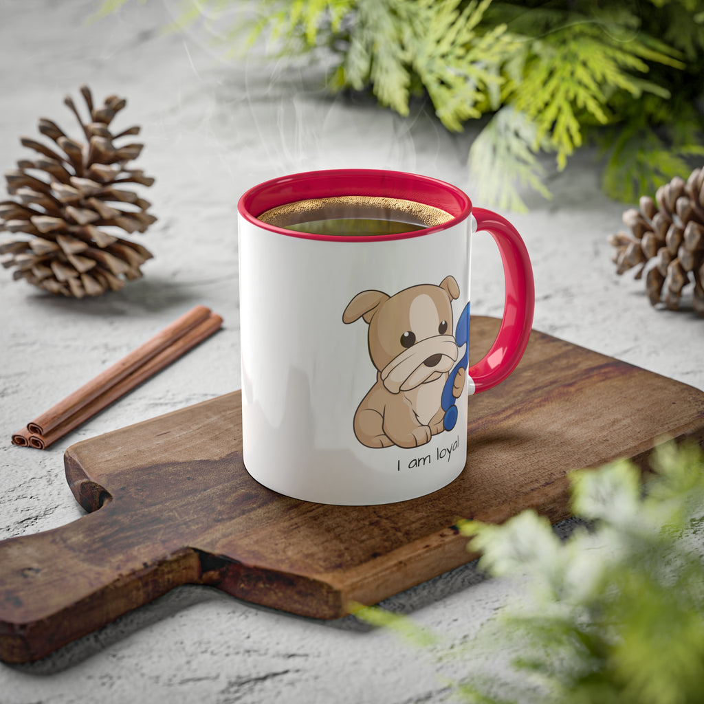 A white mug with a red handle and interior and a picture of a dog that says I am loyal. The mug has hot coffee in it and sits on a wood cutting board surrounded by pinecones and pine trees.