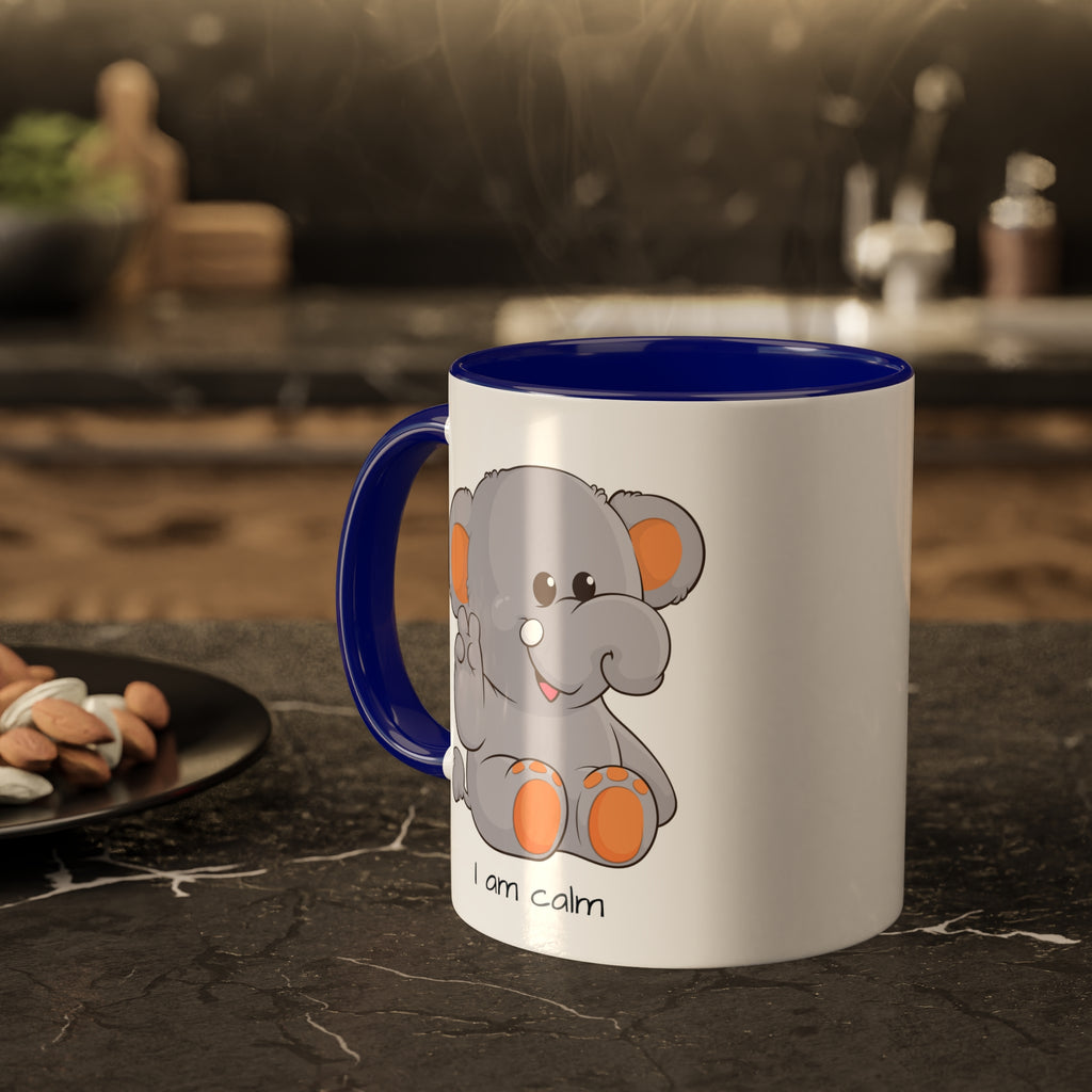 A white mug with a dark blue handle and interior and a picture of an elephant that says I am calm. The mug sits on a black kitchen counter and has steam rising out of it.