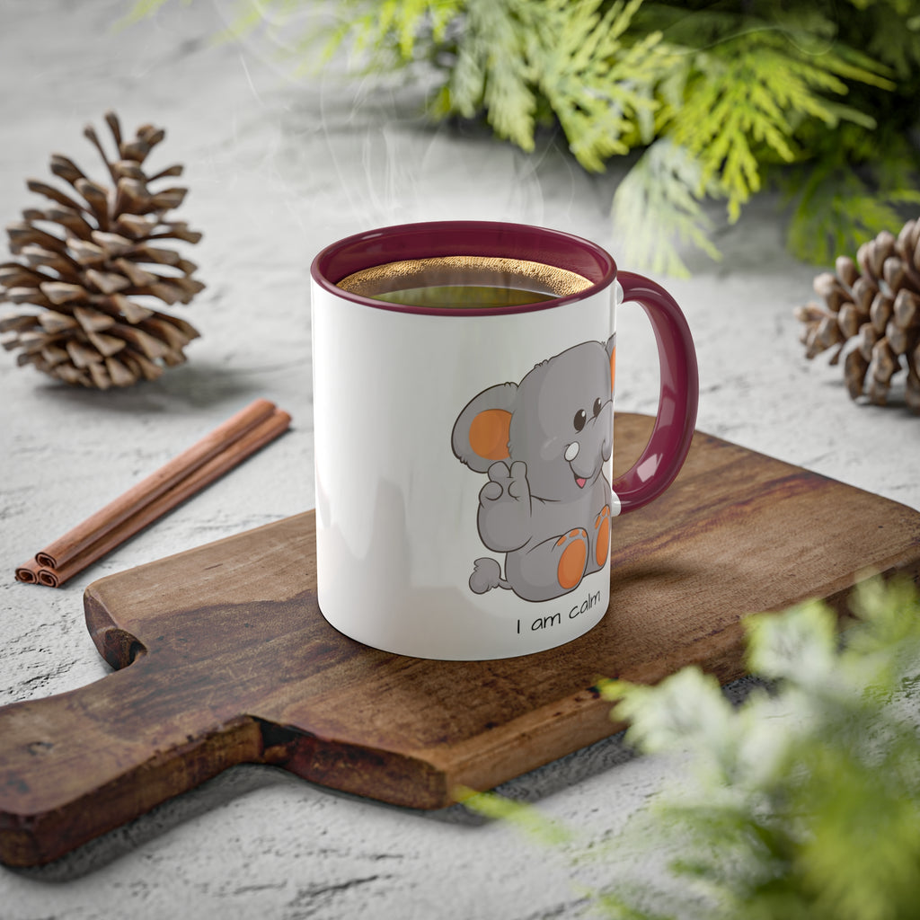 A white mug with a maroon handle and interior and a picture of an elephant that says I am calm. The mug has hot coffee in it and sits on a wood cutting board surrounded by pinecones and pine trees.