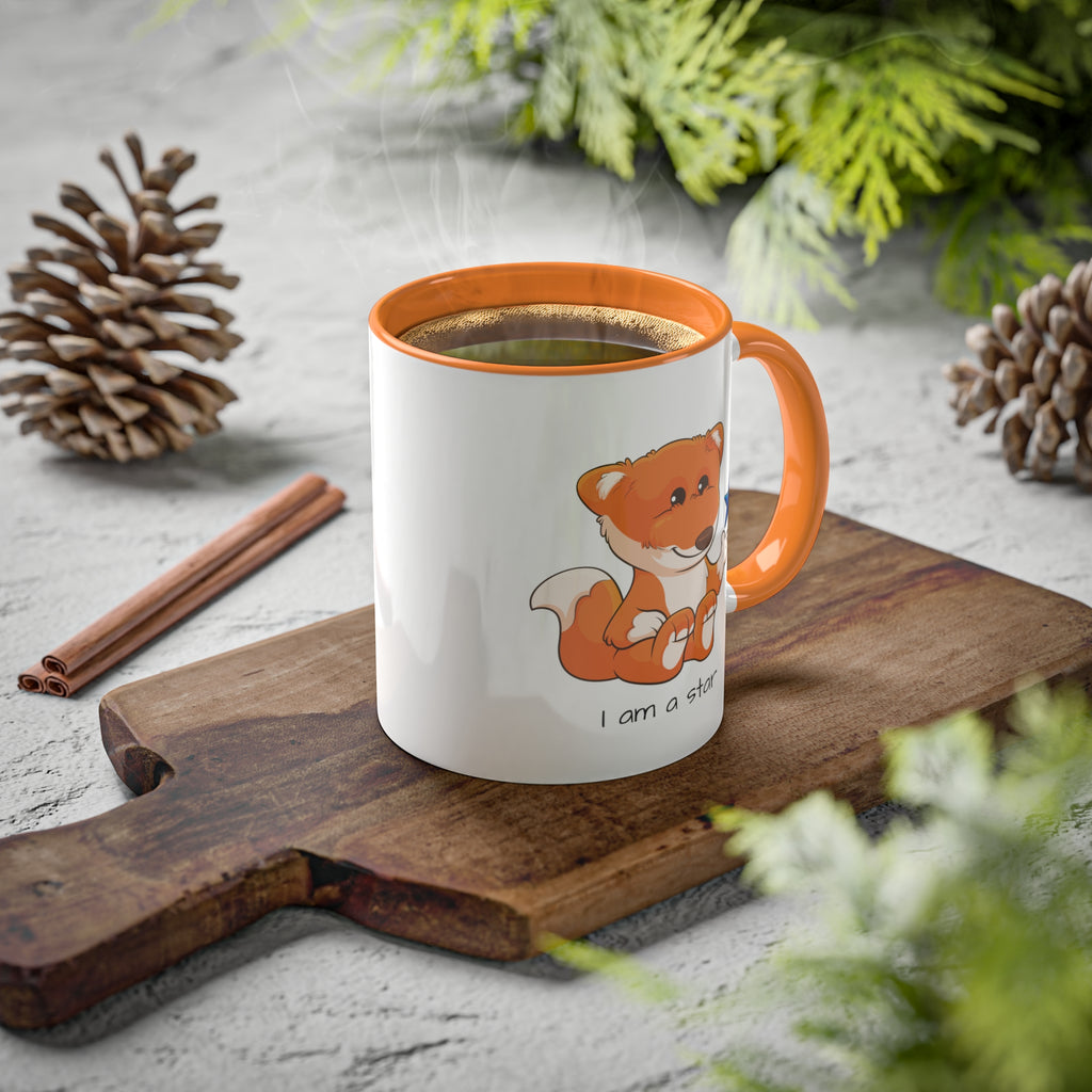 A white mug with a golden yellow handle and interior and a picture of a fox that says I am a star. The mug has hot coffee in it and sits on a wood cutting board surrounded by pinecones and pine trees.