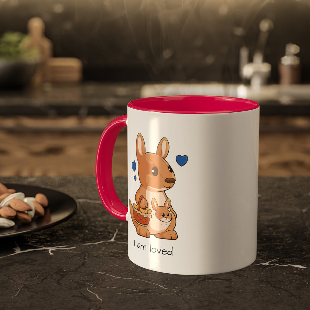 A white mug with a red handle and interior and a picture of a kangaroo that says I am loved. The mug sits on a black kitchen counter and has steam rising out of it.
