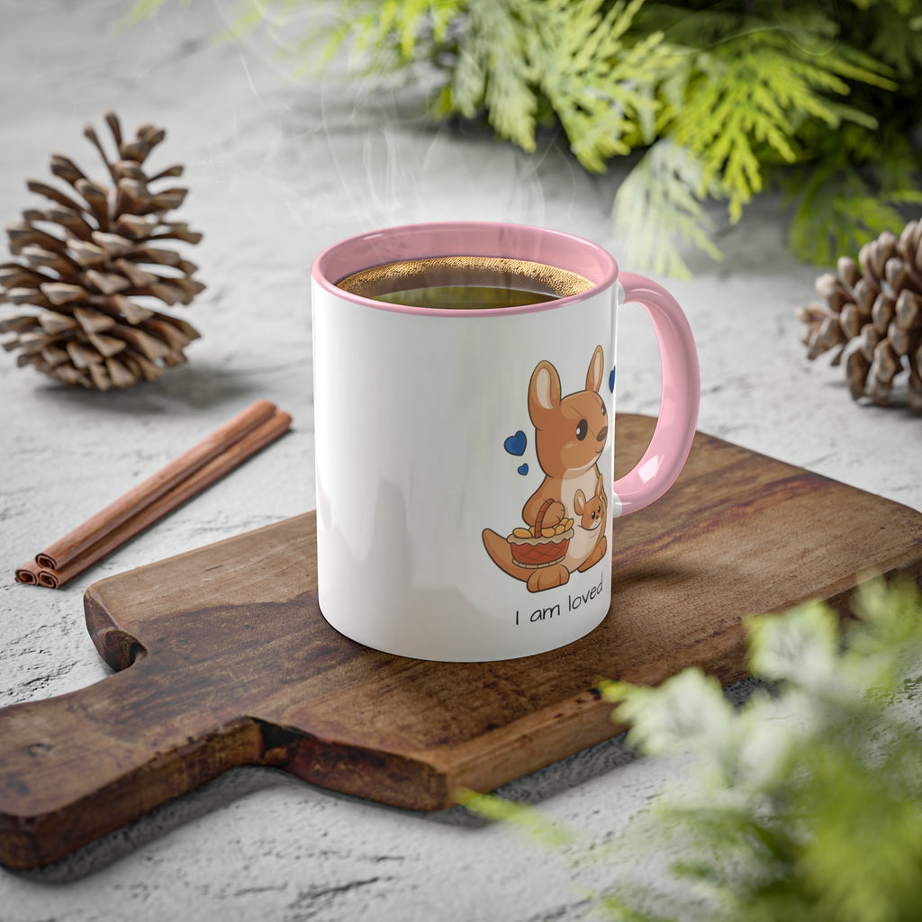 A white mug with a pink handle and interior and a picture of a kangaroo that says I am loved. The mug has hot coffee in it and sits on a wood cutting board surrounded by pinecones and pine trees.