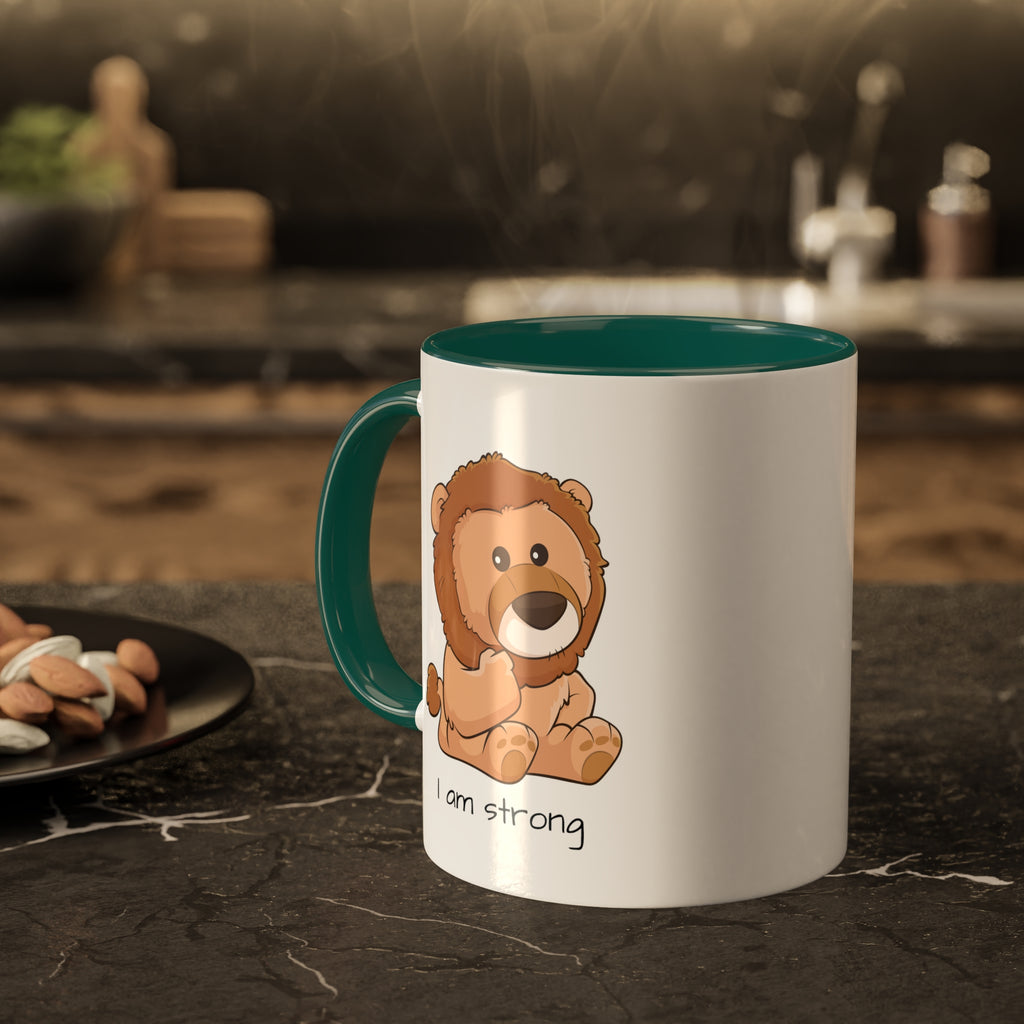A white mug with a dark green handle and interior and a picture of a lion that says I am strong. The mug sits on a black kitchen counter and has steam rising out of it.