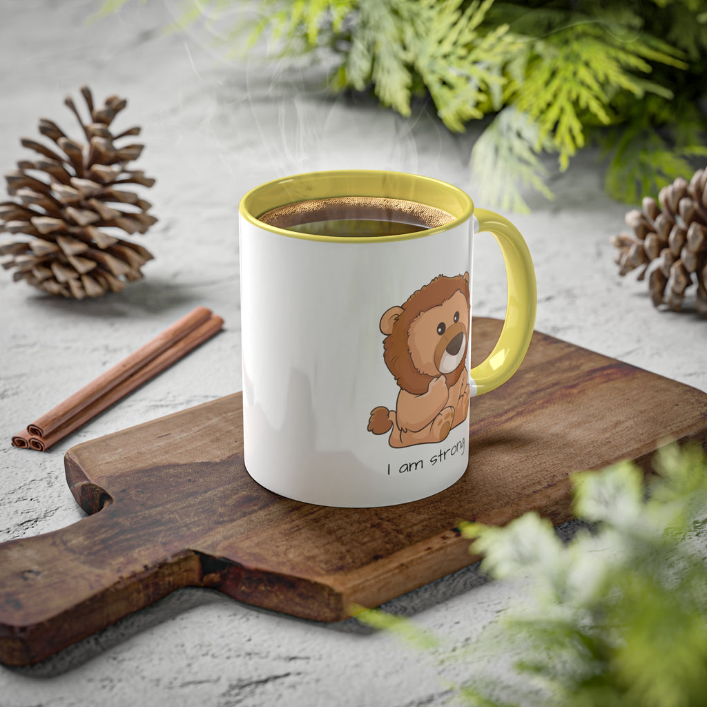 A white mug with a yellow handle and interior and a picture of a lion that says I am strong. The mug has hot coffee in it and sits on a wood cutting board surrounded by pinecones and pine trees.