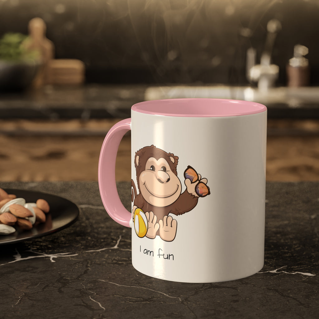 A white mug with a pink handle and interior and a picture of a monkey that says I am fun. The mug sits on a black kitchen counter and has steam rising out of it.
