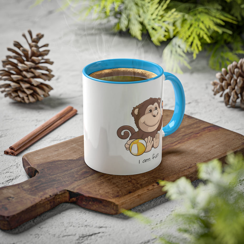 A white mug with a light blue handle and interior and a picture of a monkey that says I am fun. The mug has hot coffee in it and sits on a wood cutting board surrounded by pinecones and pine trees.