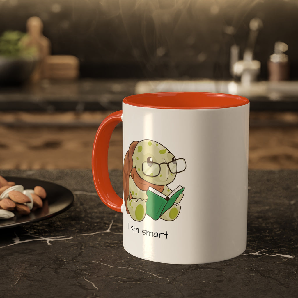 A white mug with an orange handle and interior and a picture of a turtle that says I am smart. The mug sits on a black kitchen counter and has steam rising out of it.