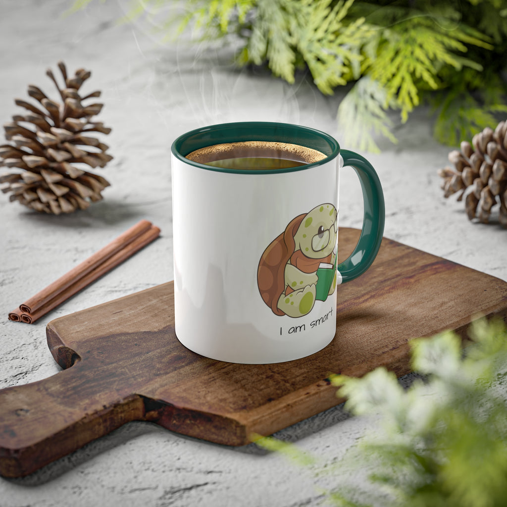 A white mug with a dark green handle and interior and a picture of a turtle that says I am smart. The mug has hot coffee in it and sits on a wood cutting board surrounded by pinecones and pine trees.